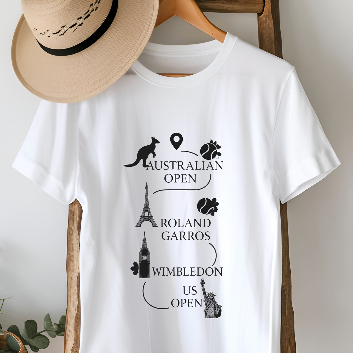 A white short-sleeve T-shirt hanging on a wooden hanger, featuring black graphic text and travel-themed illustrations including kangaroo, Eiffel Tower, Big Ben, London Eye, Statue of Liberty, and tennis pins. A beige straw hat is draped over the collar, and part of a wooden chair is visible in a styled indoor setting.